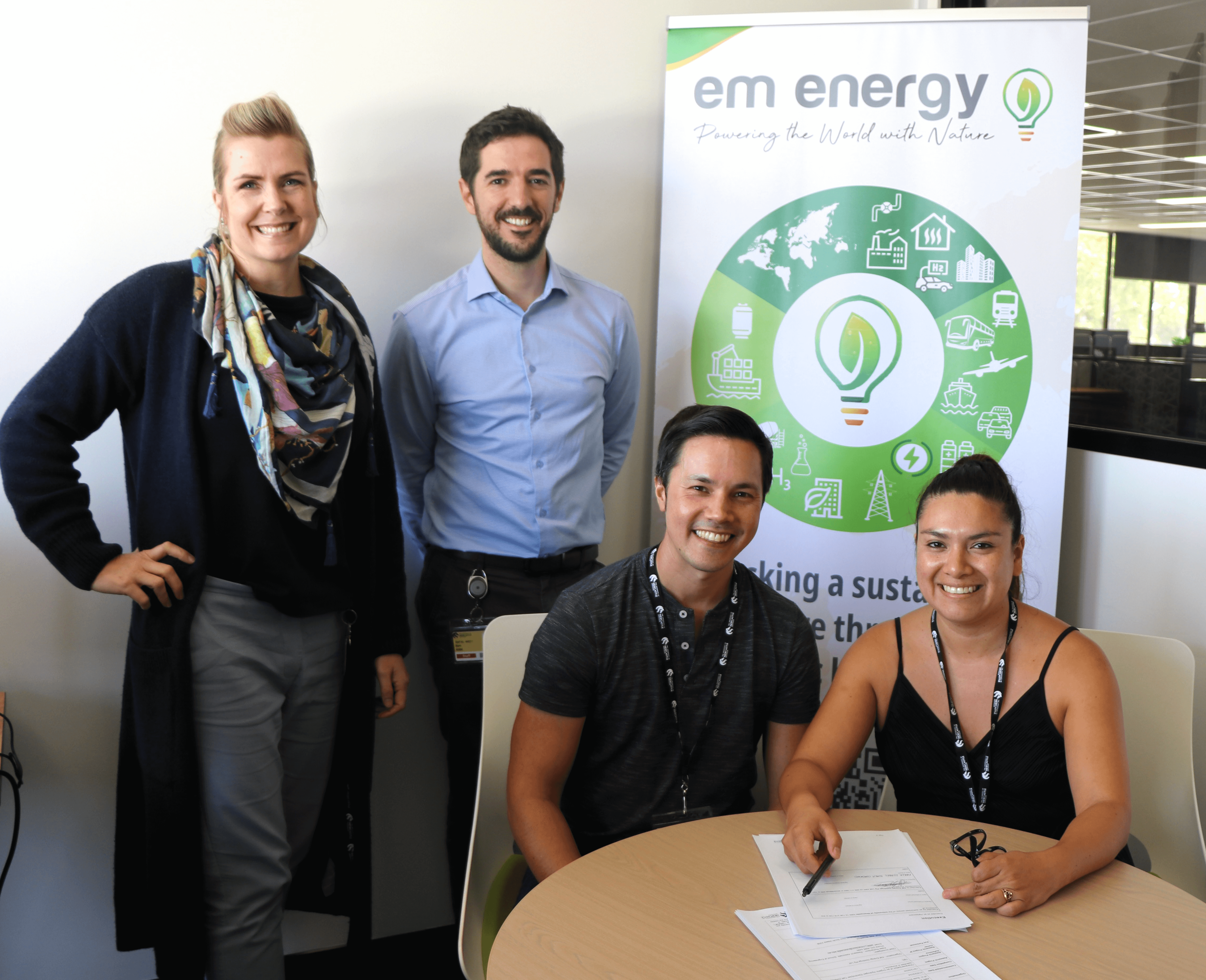 EM Energy sitting at a desk about to sign their R&D voucher paperwork. TRaCE team members are standing next to them. Everyone is smiling at the camera.