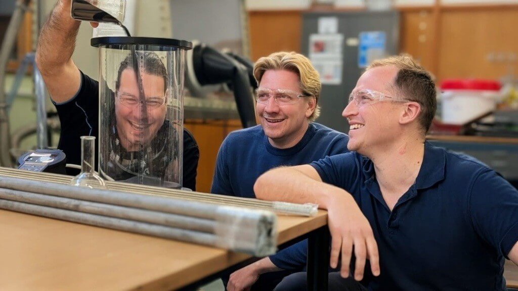 Three men wearing safety glasses, sitting at a table. The man on the left is pouring particles into a cylindrical glass container while the other two watch.